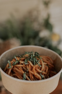 A biodegradable bowl filled with saucy noodles garnished with chopped herbs, set against a softly blurred background that likely includes plants.