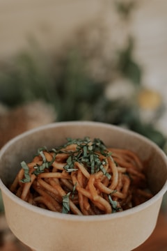 A biodegradable bowl filled with saucy noodles garnished with chopped herbs, set against a softly blurred background that likely includes plants.