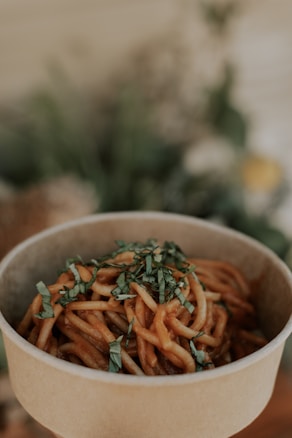 A biodegradable bowl filled with saucy noodles garnished with chopped herbs, set against a softly blurred background that likely includes plants.
