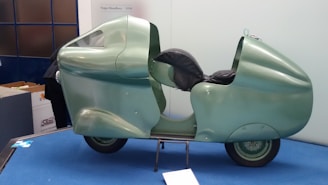 A uniquely designed, streamlined motorcycle displayed on a blue platform. Its body is covered in a smooth, shiny light green casing with aerodynamic curves and a black leather seat. The setting suggests a museum or exhibition environment.