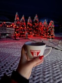 A hand holding a mug with a football club logo in the foreground. The background has snow-covered trees adorned with red lights creating a festive atmosphere.