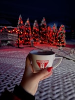 A hand holding a mug with a football club logo in the foreground. The background has snow-covered trees adorned with red lights creating a festive atmosphere.