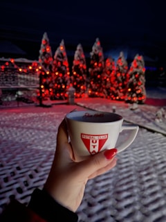 A hand holding a mug with a football club logo in the foreground. The background has snow-covered trees adorned with red lights creating a festive atmosphere.