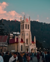 A large crowd gathers in front of a historic Gothic-style church with a prominent red cross on its steeple. The building is cream-colored with a red roof, set against a backdrop of dense green forest under a sky with scattered clouds. There are several people in winter clothing, indicating a chilly setting.