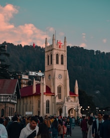 A large crowd gathers in front of a historic Gothic-style church with a prominent red cross on its steeple. The building is cream-colored with a red roof, set against a backdrop of dense green forest under a sky with scattered clouds. There are several people in winter clothing, indicating a chilly setting.