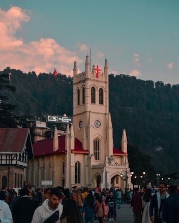 A large crowd gathers in front of a historic Gothic-style church with a prominent red cross on its steeple. The building is cream-colored with a red roof, set against a backdrop of dense green forest under a sky with scattered clouds. There are several people in winter clothing, indicating a chilly setting.
