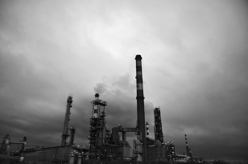 A large industrial facility with several tall chimneys and complex metal structures is set against a cloudy sky. The atmosphere appears overcast, enhancing the stark industrial aesthetic.