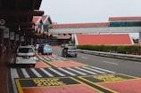 A busy airport drop-off area with multiple vehicles parked and passing by. There are red and yellow marked zones indicating 'DROP ZONE' on the pavement, and a modern terminal building with red roofs in the background. Signage and traffic lines guide the flow of cars.