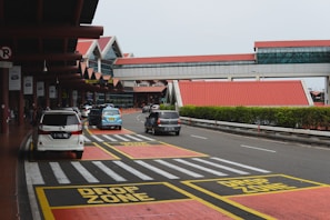 A busy airport drop-off area with multiple vehicles parked and passing by. There are red and yellow marked zones indicating 'DROP ZONE' on the pavement, and a modern terminal building with red roofs in the background. Signage and traffic lines guide the flow of cars.