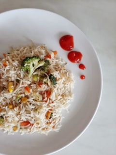A plate of colorful vegetable fried rice containing broccoli, corn, and carrots is garnished with three dollops of ketchup on a white dish. The rice is mixed with a variety of vegetables, providing a vibrant contrast.