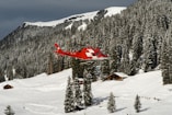 a red helicopter flying over a snow covered mountain
