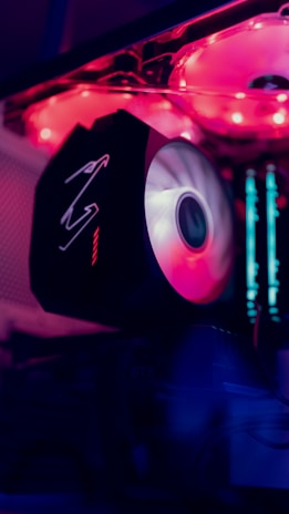 A close-up of a gaming PC build featuring a glowing LED fan with a prominent brand logo. The inside of the case emits a vibrant pink and purple illumination with visible components such as RAM sticks and cooling fans.