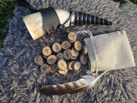 A rustic setting featuring a drinking horn with a black strap and spiral tip, a collection of wooden rune discs with carvings, a cloth drawstring pouch, and a feather. These items rest on a textured, fluffy gray surface, possibly faux fur or fleece.