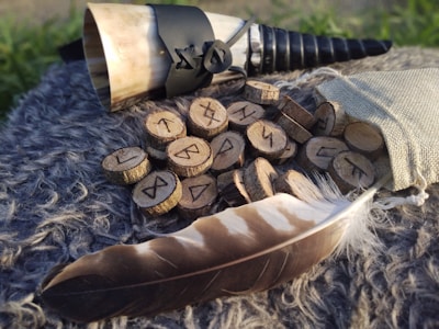 A collection of wooden discs with carved runes is scattered on a fur surface. A horn adorned with black leather accents and a rope lacing is placed beside them. A faded burlap pouch lies nearby, with some runes spilling out. A large, brown and white feather is also visible in the foreground.