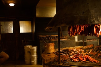 A rustic indoor barbecue setting with sausages hanging above a large grill filled with various meats. The room is dimly lit with a warm glow, casting shadows on wooden walls and surrounding objects, including buckets and cooking utensils.