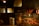 A rustic indoor barbecue setting with sausages hanging above a large grill filled with various meats. The room is dimly lit with a warm glow, casting shadows on wooden walls and surrounding objects, including buckets and cooking utensils.