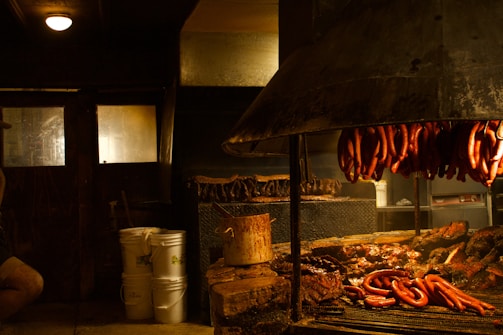A rustic indoor barbecue setting with sausages hanging above a large grill filled with various meats. The room is dimly lit with a warm glow, casting shadows on wooden walls and surrounding objects, including buckets and cooking utensils.