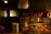 A rustic indoor barbecue setting with sausages hanging above a large grill filled with various meats. The room is dimly lit with a warm glow, casting shadows on wooden walls and surrounding objects, including buckets and cooking utensils.