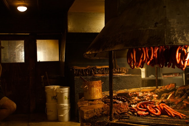 A rustic indoor barbecue setting with sausages hanging above a large grill filled with various meats. The room is dimly lit with a warm glow, casting shadows on wooden walls and surrounding objects, including buckets and cooking utensils.