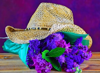 A straw cowboy hat with a decorative band is placed on top of a bouquet of vibrant purple flowers, wrapped in green paper. The hat and flowers rest on a wooden surface, with a blurred purple background adding depth.