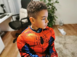 A young boy wearing a red and blue superhero costume with a black spider emblem is standing in an indoor setting. The room contains an office chair, a desk, and a green plant. The boy is looking to the side with a focused expression.