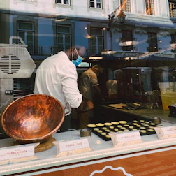 A person wearing a mask works inside a pastry shop, preparing pastries on a countertop. A large copper bowl is visible in the foreground, and several pastries are arranged in rows on baking trays. The shop is identified as Fábrica de Nata, and the interior is modern with reflections of the street visible on the glass window.