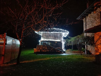 A small rustic building with a tiled roof is illuminated by bright string lights. It sits in a grassy area surrounded by trees, with a stone wall in front. The surroundings are dark, indicating night time, and there is another building partially visible on the right side.