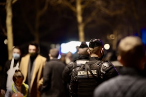 A group of people stands in an outdoor setting, with a uniformed officer in focus wearing a vest labeled 'Gendarmerie'. The scene is dimly lit and the background is blurred, suggesting a night-time or evening environment.