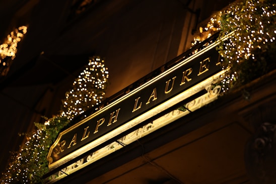 An illuminated store sign displaying 'Ralph Lauren' in elegant gold lettering is surrounded by decorative lights and greenery. The warm glow from the lights creates a luxurious and inviting atmosphere.