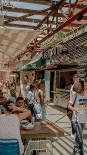 A lively outdoor market scene with several people casually interacting and lining up at a food stall. The area is covered by a pergola with string lights, creating a shaded, relaxed atmosphere. Various signs and restaurant names are visible, adding to the bustling and vibrant setting.