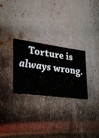 A bold, rectangular black sign on a textured concrete wall with the text 'Torture is always wrong' printed in white, conveying a strong anti-torture message.