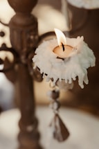 A lit candle with melted wax drips hanging from the sides, placed on an ornate, antique-looking holder. The flame is small, casting a gentle light and is surrounded by intricate metalwork and a blurred background.