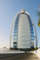 A tall, sail-shaped luxury hotel with a unique architectural design stands under a clear blue sky. The building is surrounded by palm trees and a fenced road leading to the entrance.