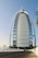 A tall, sail-shaped luxury hotel with a unique architectural design stands under a clear blue sky. The building is surrounded by palm trees and a fenced road leading to the entrance.
