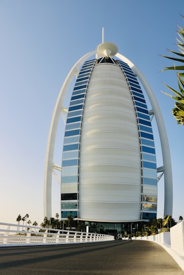 A tall, sail-shaped luxury hotel with a unique architectural design stands under a clear blue sky. The building is surrounded by palm trees and a fenced road leading to the entrance.