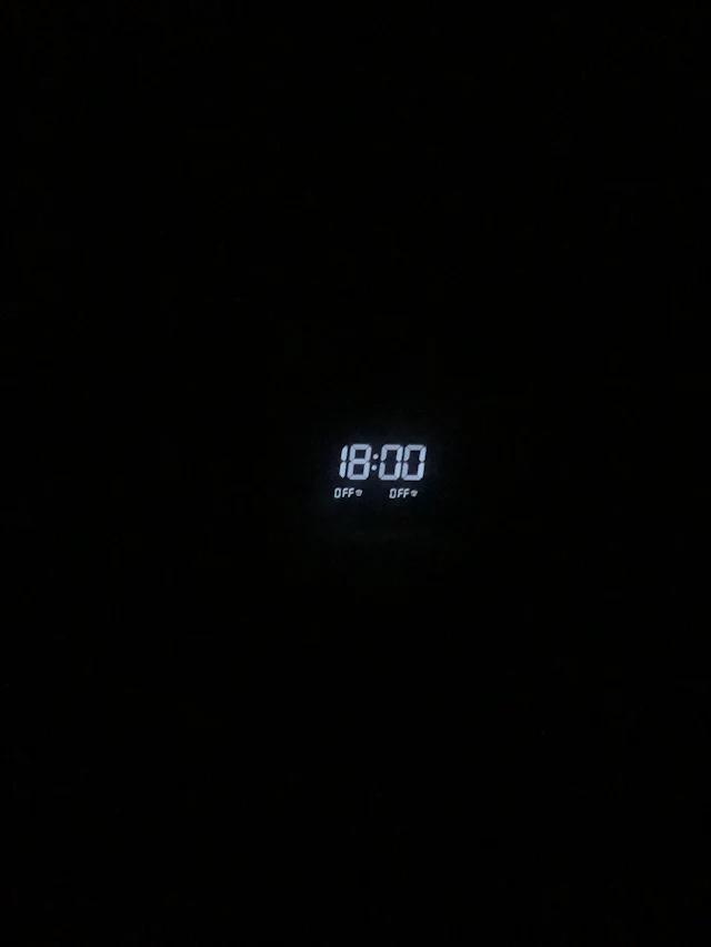 Relógio digital de LED branco mostrando 18:00 em um ambiente escuro.