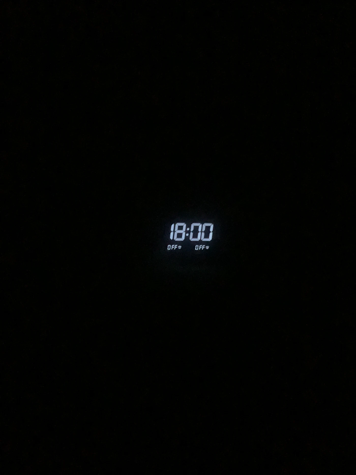 Relógio digital de LED branco mostrando 18:00 em um ambiente escuro.