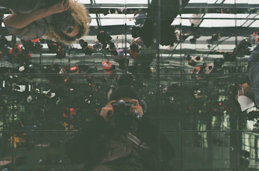 A complex reflection scene featuring multiple people looking at their own reflections in a mirrored ceiling. The image captures numerous reflections creating an intricate, kaleidoscope-like effect. Individuals are positioned at various angles, with a prominent figure holding a camera, possibly a photographer capturing this surreal moment. The scene is bustling with activity, as seen in the various figures scattered across the reflective surface.