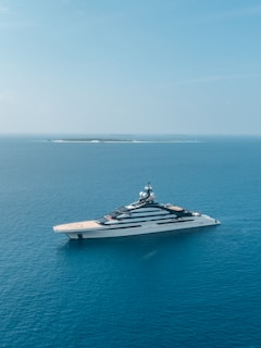 A luxurious yacht is sailing on calm, clear blue ocean waters. The backdrop features a distant island with lush greenery and minimal structures. The serene sky is a light blue, blending harmoniously with the sea.