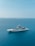A luxurious yacht is sailing on calm, clear blue ocean waters. The backdrop features a distant island with lush greenery and minimal structures. The serene sky is a light blue, blending harmoniously with the sea.