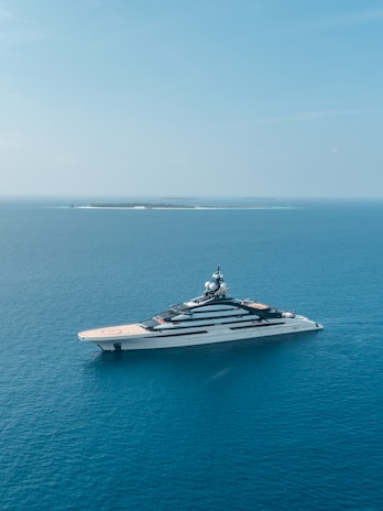 A luxurious yacht is sailing on calm, clear blue ocean waters. The backdrop features a distant island with lush greenery and minimal structures. The serene sky is a light blue, blending harmoniously with the sea.