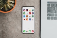 A sleek smartphone displaying various banking app icons is placed on a grey surface. To the left, there is a potted succulent plant, and on the right, part of a laptop keyboard is visible.