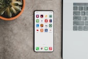 A sleek smartphone displaying various banking app icons is placed on a grey surface. To the left, there is a potted succulent plant, and on the right, part of a laptop keyboard is visible.