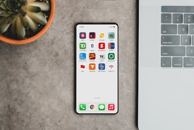 A sleek smartphone displaying various banking app icons is placed on a grey surface. To the left, there is a potted succulent plant, and on the right, part of a laptop keyboard is visible.