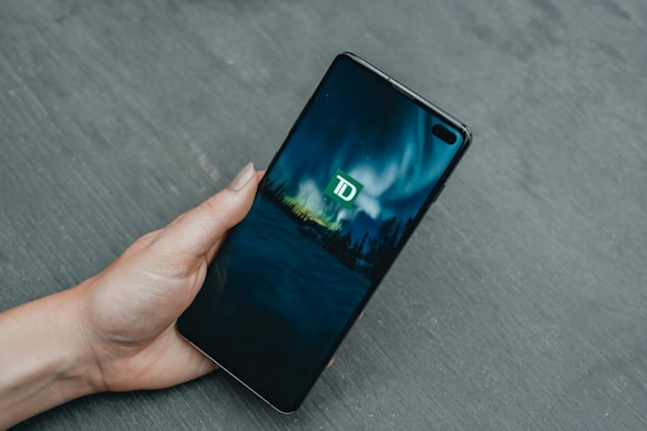 A hand holds a smartphone displaying a screen with the TD logo against a backdrop of a stylized aurora borealis. The phone is angled slightly, revealing the front camera and sensors.