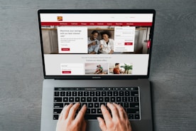 A person is using a MacBook Pro to browse a banking website. The screen displays a login section, promotional offers, and an image of a smiling couple in the top section. The design includes a red color scheme, with navigation links for various banking services.