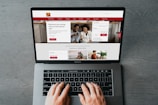 A person is using a MacBook Pro to browse a banking website. The screen displays a login section, promotional offers, and an image of a smiling couple in the top section. The design includes a red color scheme, with navigation links for various banking services.