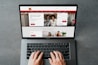 A person is using a MacBook Pro to browse a banking website. The screen displays a login section, promotional offers, and an image of a smiling couple in the top section. The design includes a red color scheme, with navigation links for various banking services.