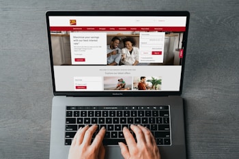 A person is using a MacBook Pro to browse a banking website. The screen displays a login section, promotional offers, and an image of a smiling couple in the top section. The design includes a red color scheme, with navigation links for various banking services.