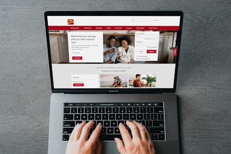 A person is using a MacBook Pro to browse a banking website. The screen displays a login section, promotional offers, and an image of a smiling couple in the top section. The design includes a red color scheme, with navigation links for various banking services.